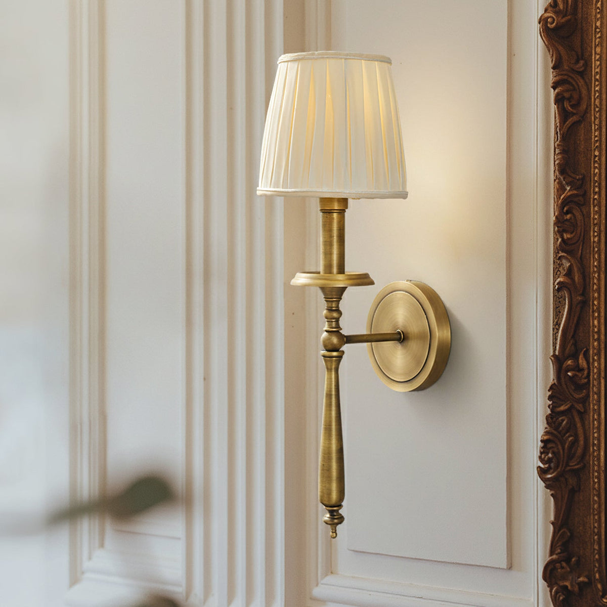 Close view of the pleated shade, turned brass stem and round backplate of this Clérachic sconce, beside an ornate dark frame.
