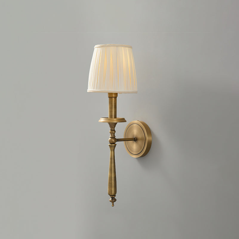 Antique brass Clérachic wall light with a pleated beige shade, shown unlit against a mid-grey wall.