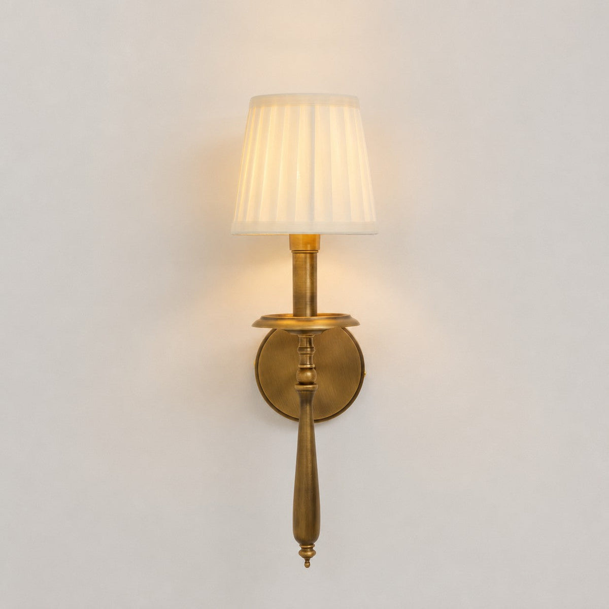 Lit Clérachic wall sconce facing front, with a warm glow through its pleated shade and a round antique brass backplate.