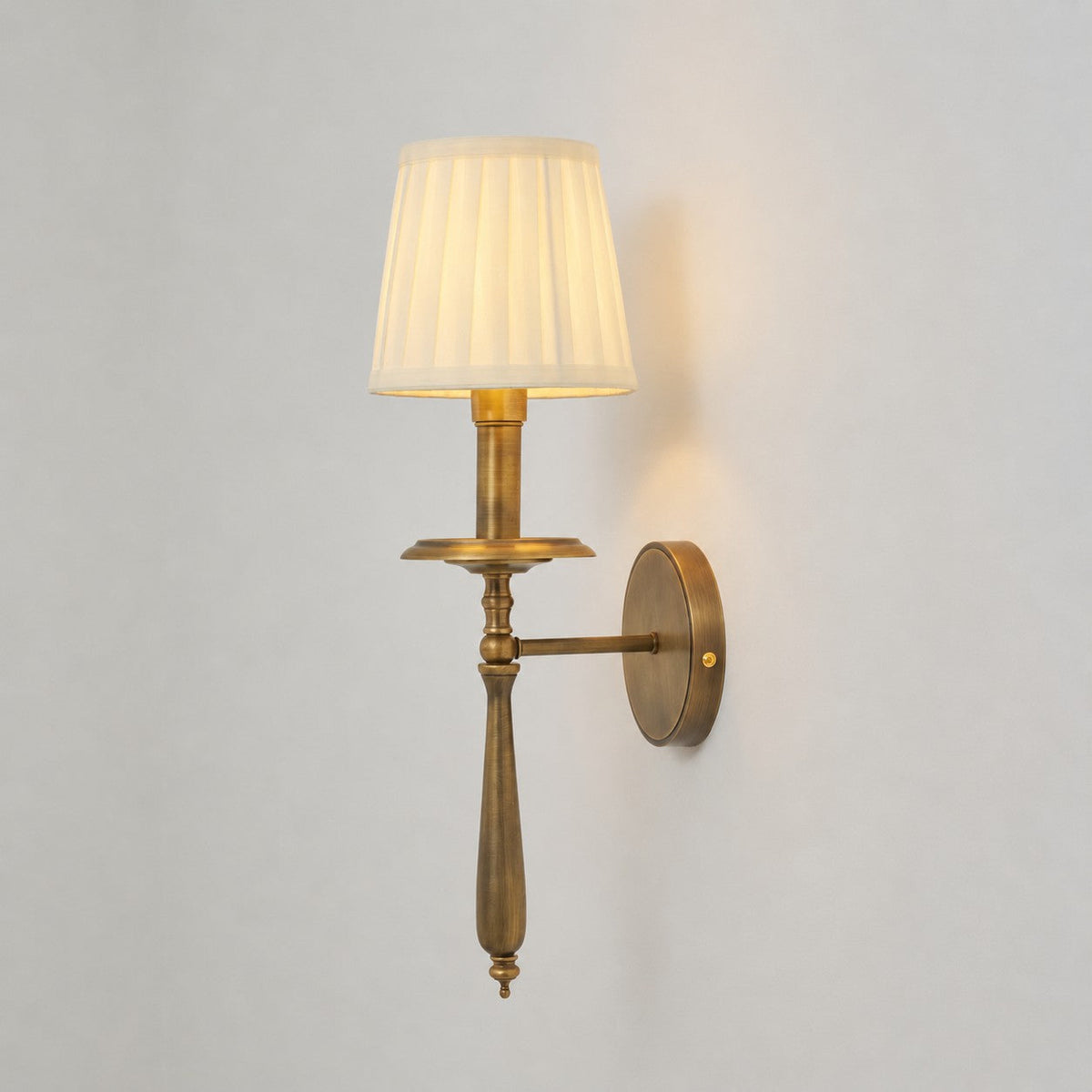 Clérachic brass wall light with a softly glowing pleated shade and a side arm to a round backplate, on a pale wall.