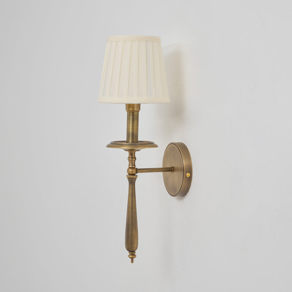 An unlit Clérachic wall sconce in antique brass with a cream pleated shade, with its stem and drip tray in clear view.