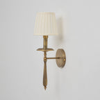 An unlit Clérachic wall sconce in antique brass with a cream pleated shade, with its stem and drip tray in clear view.