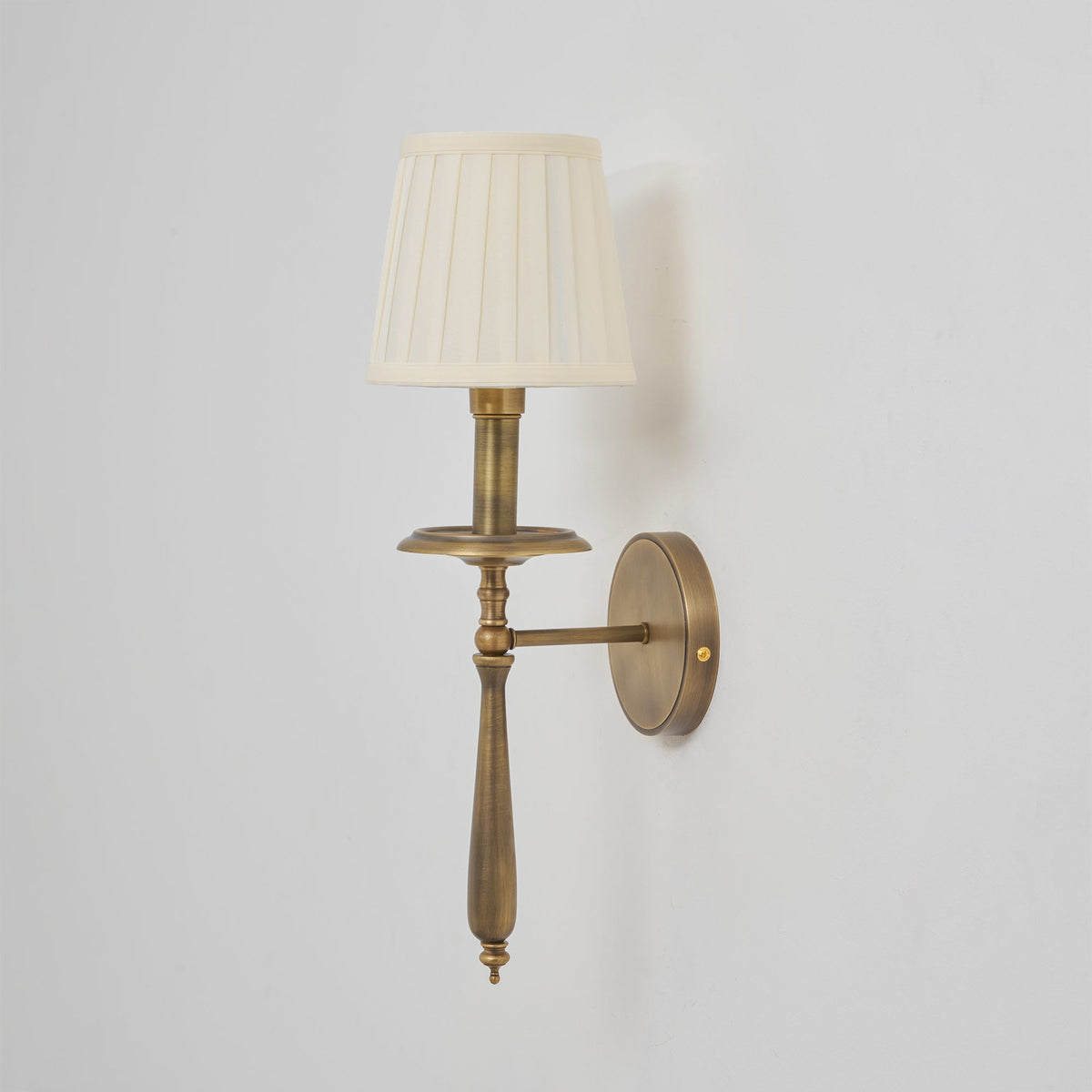 An unlit Clérachic wall sconce in antique brass with a cream pleated shade, with its stem and drip tray in clear view.