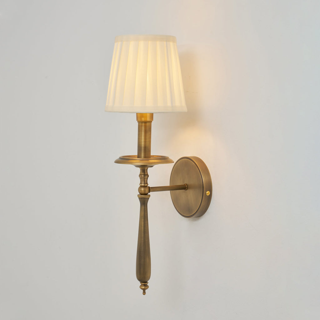 Clérachic wall sconce with a lit pleated shade, a brass drip tray and a turned stem, on a pale grey wall.