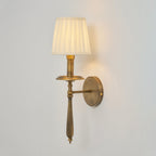 Clérachic wall sconce with a lit pleated shade, a brass drip tray and a turned stem, on a pale grey wall.