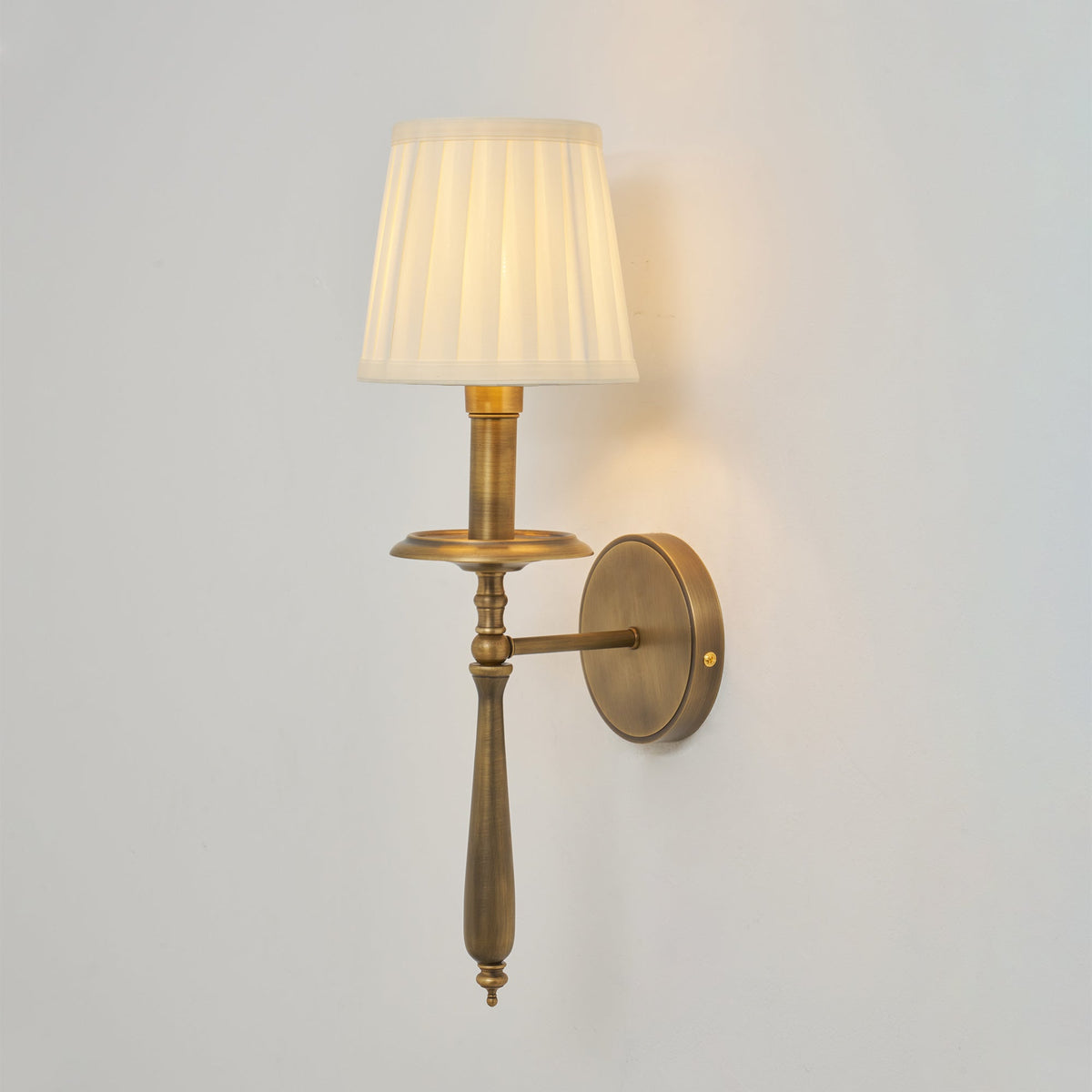 Clérachic wall sconce with a lit pleated shade, a brass drip tray and a turned stem, on a pale grey wall.