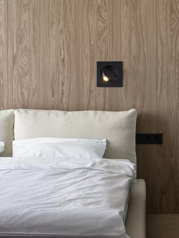 A small black Clérachic recessed wall light glows on a wood-veneer wall above a bed with white linen and a cream headboard.