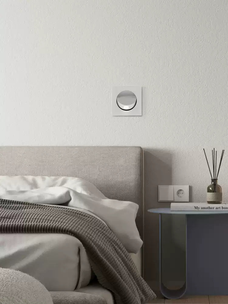 This Clérachic recessed wall light in a white finish sits on a pale wall above a bed with a grey throw and a bedside table.