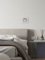 This Clérachic recessed wall light in a white finish sits on a pale wall above a bed with a grey throw and a bedside table.