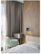 Wide view of a black Clérachic recessed wall light in a wood-panelled wall, next to full-length curtains and a made bed.