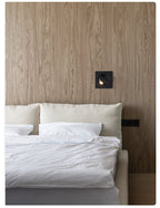 Black Clérachic recessed wall light set into a wood-grain wall above the headboard of a bed with white bedding.