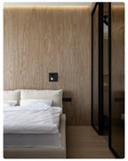 A small lit Clérachic recessed wall light on a wood-veneer wall beside a dark glass door and a bed with white linen.