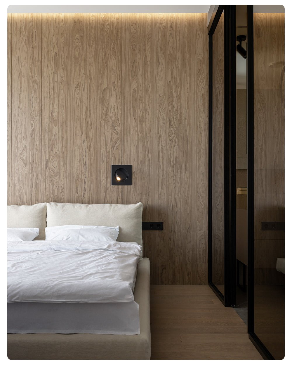 A small lit Clérachic recessed wall light on a wood-veneer wall beside a dark glass door and a bed with white linen.