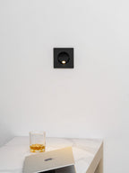 Lit black Clérachic recessed wall light on a white wall above a marble-topped desk with a whisky glass and a laptop.