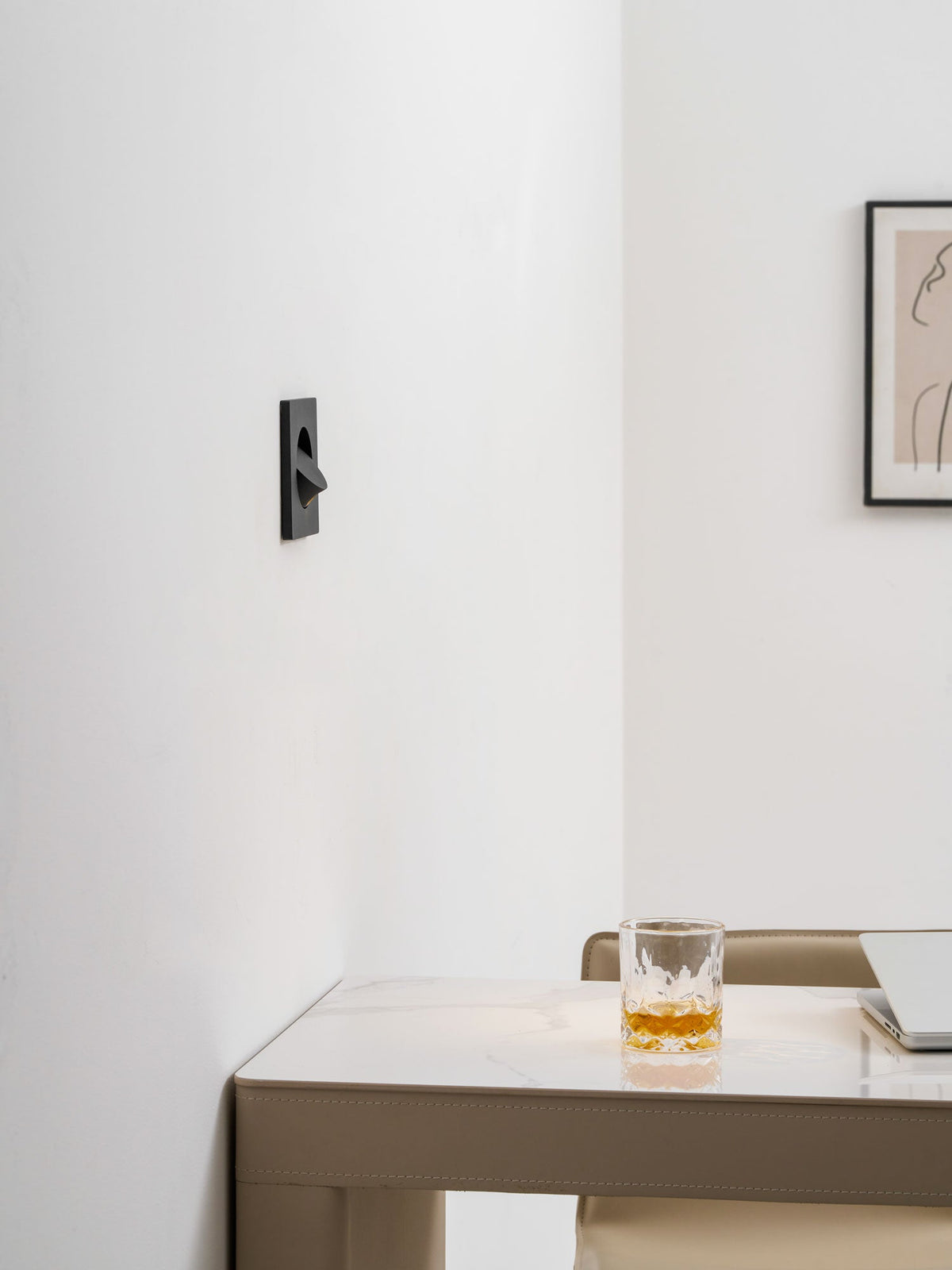 Side-on view of this Clérachic recessed wall light with its hood turned, on a white wall above a marble desk and a glass.