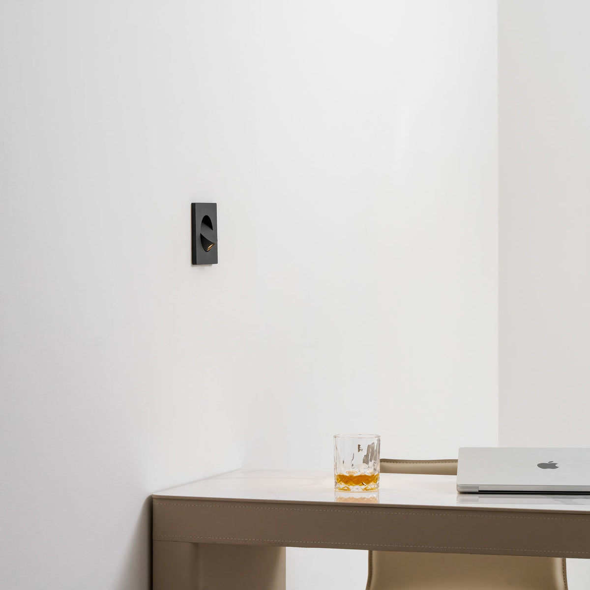 The black Clérachic recessed wall light seen from the side on a white wall, with a glass and a laptop on the desk below.
