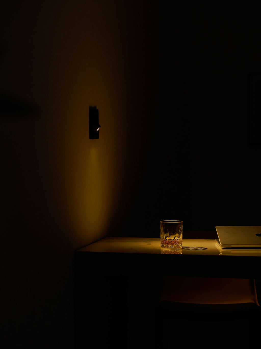 In a dark room, a Clérachic recessed wall light gives off a faint warm glow over a desk, a glass and a laptop.