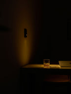 In a dark room, a Clérachic recessed wall light gives off a faint warm glow over a desk, a glass and a laptop.