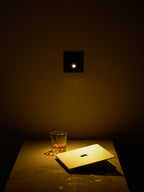A single warm LED glows from the square black Clérachic wall light, lighting a desk, a glass and an open laptop below.