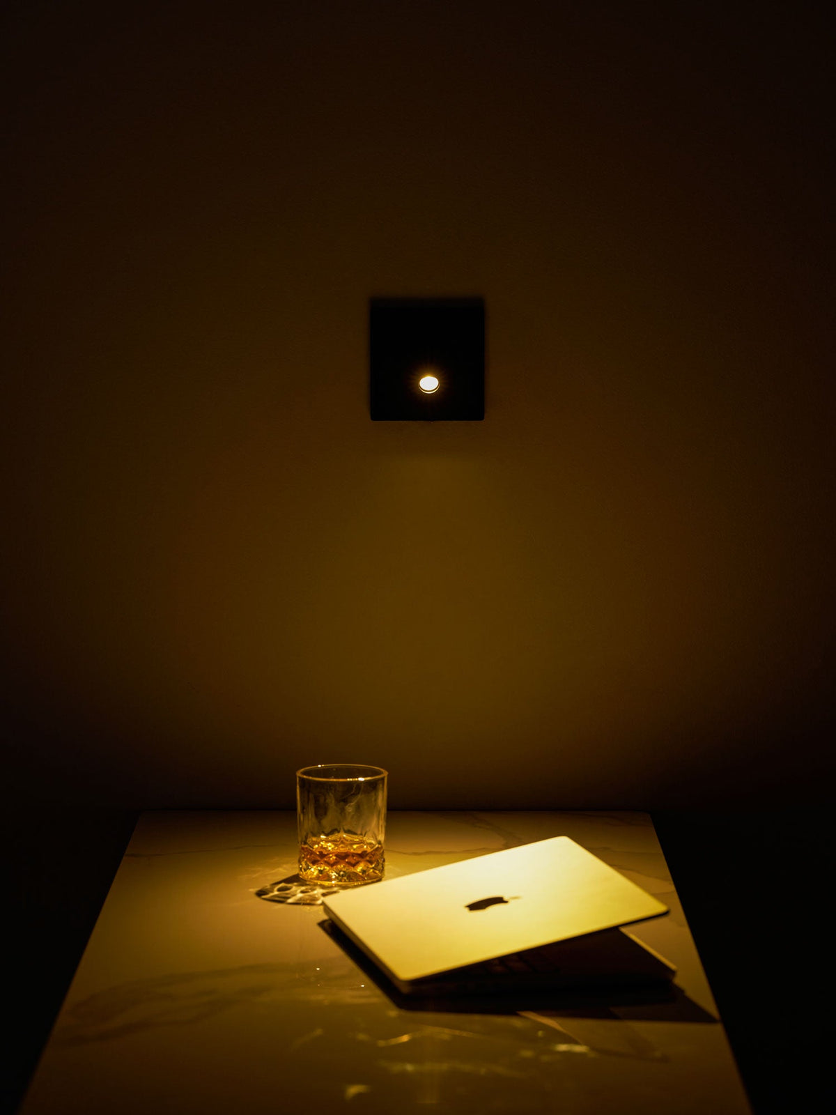 A single warm LED glows from the square black Clérachic wall light, lighting a desk, a glass and an open laptop below.
