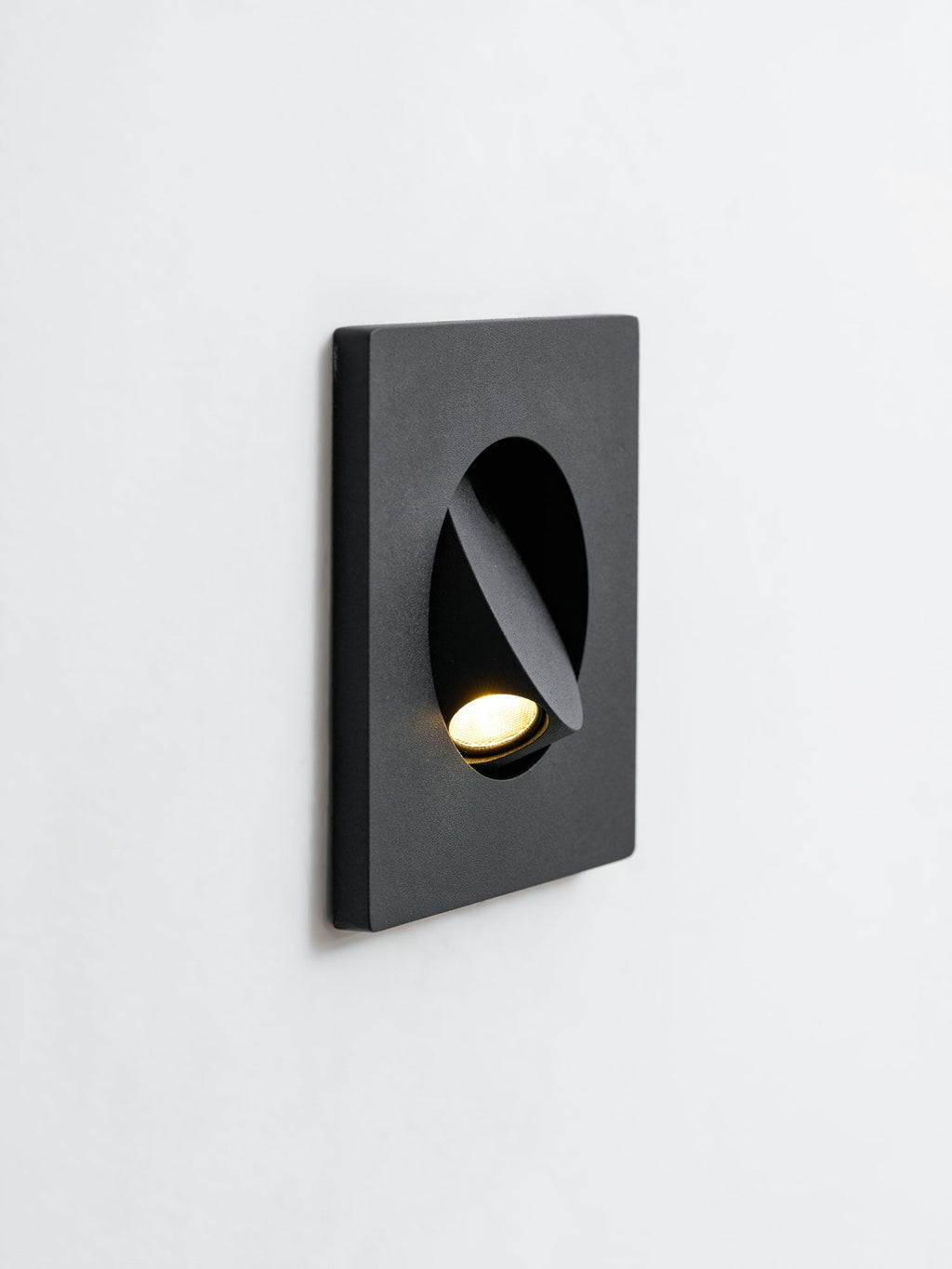 Angled close view of a black Clérachic recessed wall light on a white wall, its hood turned to aim the warm LED downward.
