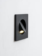 Angled close view of a black Clérachic recessed wall light on a white wall, its hood turned to aim the warm LED downward.