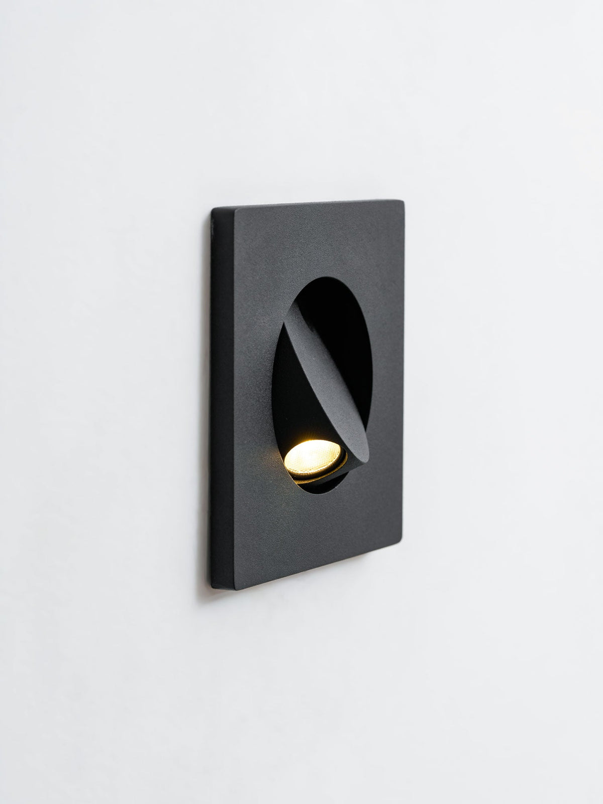 Angled close view of a black Clérachic recessed wall light on a white wall, its hood turned to aim the warm LED downward.
