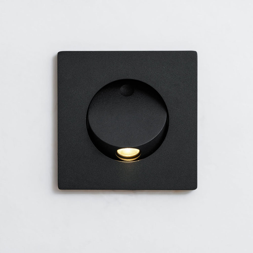 Front close view of the square black Clérachic recessed wall light, with its round opening, hood and warm LED lit.