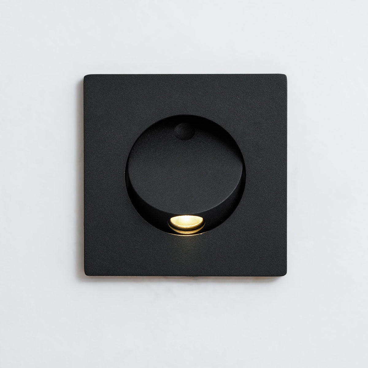 Front close view of the square black Clérachic recessed wall light, with its round opening, hood and warm LED lit.