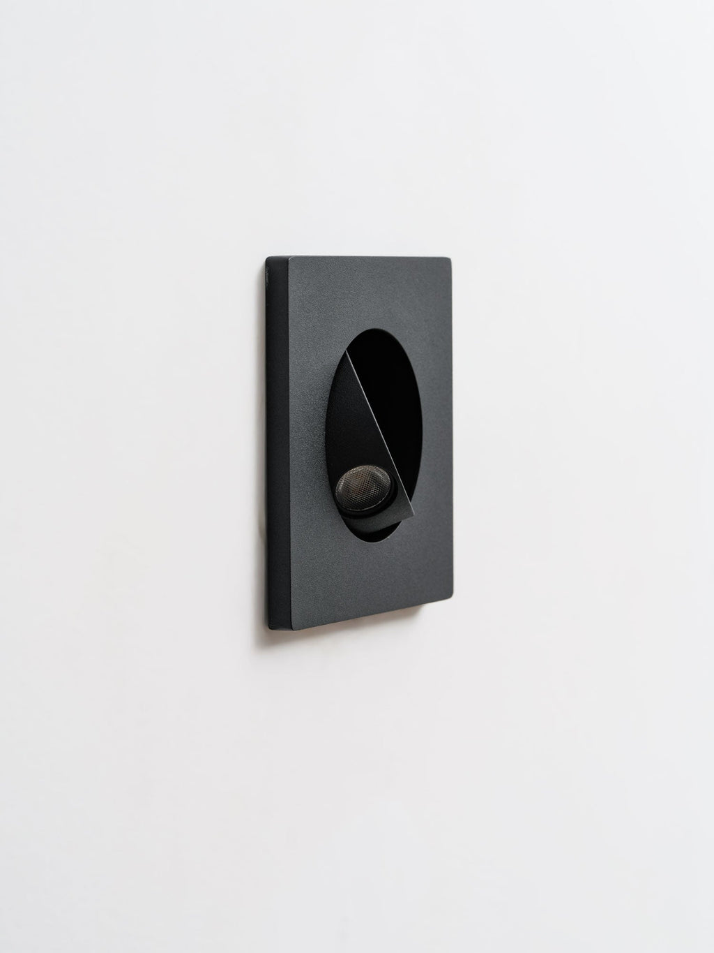 Angled view of a black Clérachic recessed wall light with its hood tilted, shown unlit on a pale wall.