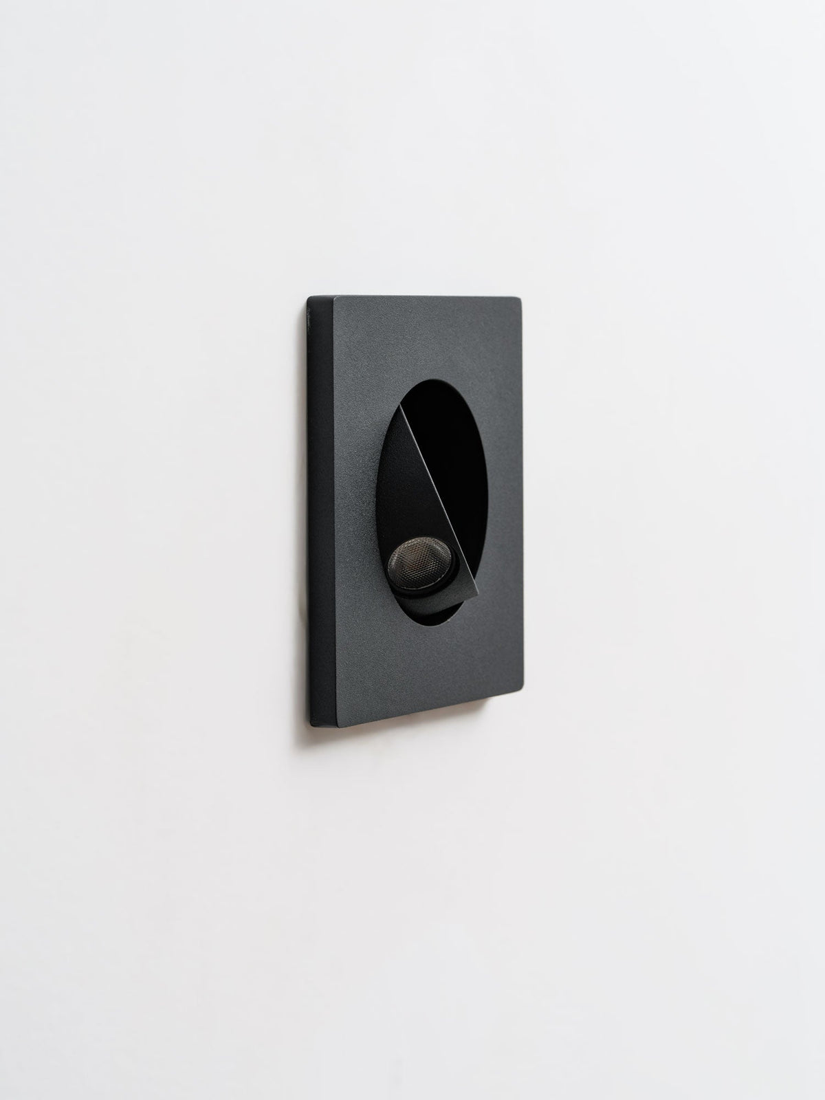 Angled view of a black Clérachic recessed wall light with its hood tilted, shown unlit on a pale wall.