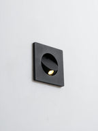 Black Clérachic recessed wall light on a pale wall, its hood tilted and the warm LED just visible in the opening.