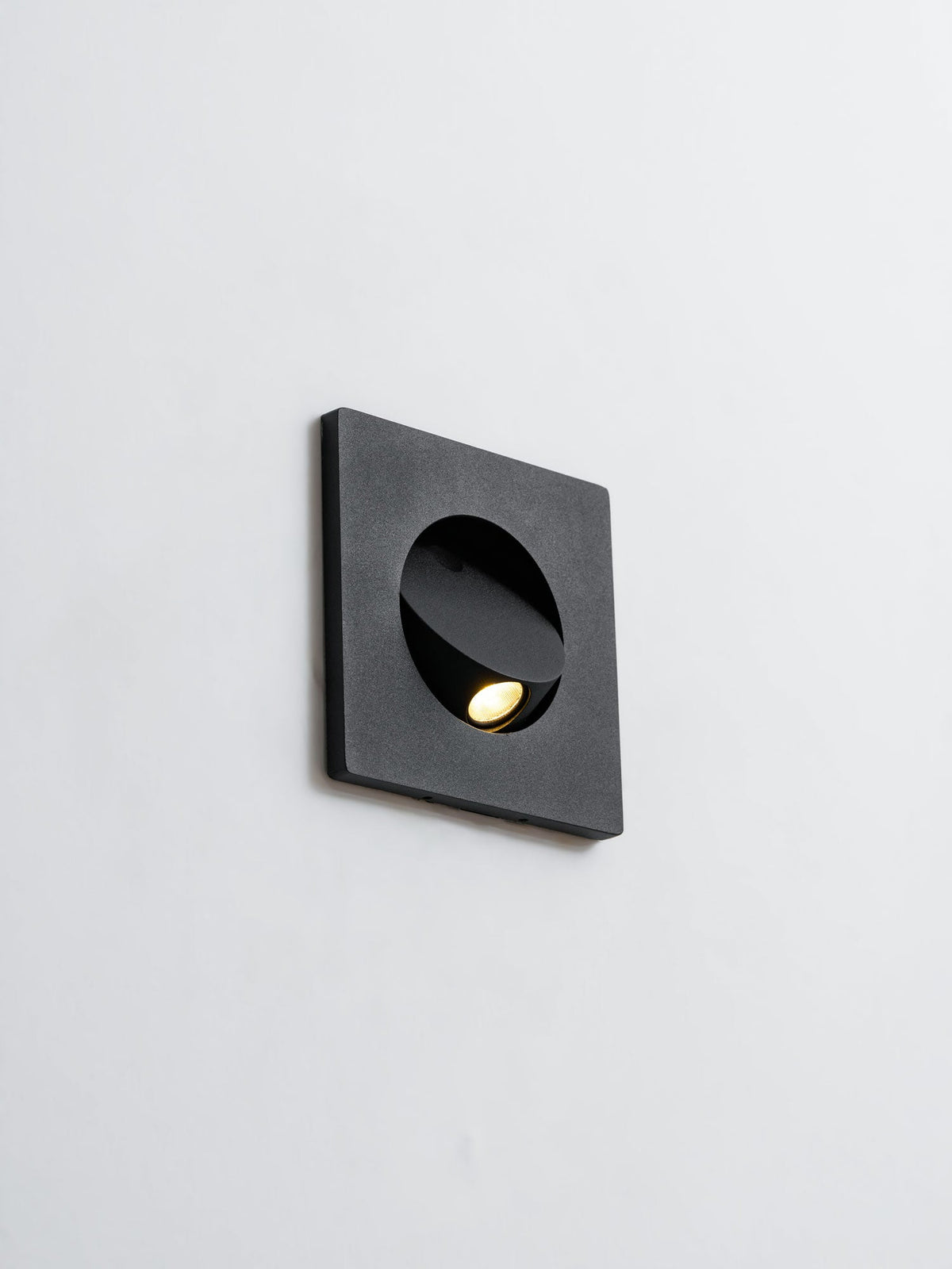 Black Clérachic recessed wall light on a pale wall, its hood tilted and the warm LED just visible in the opening.