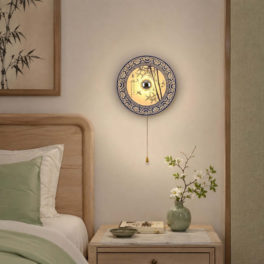 A Clérachic wall light with a bamboo-pattern porcelain shade above a bedside table, beside a sage-green pillow and a small vase of blossom.