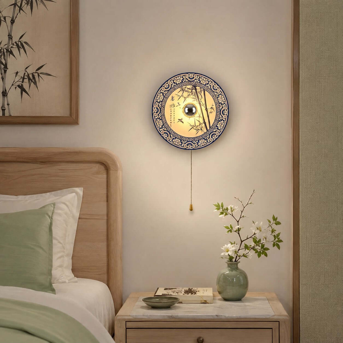 A Clérachic wall light with a bamboo-pattern porcelain shade above a bedside table, beside a sage-green pillow and a small vase of blossom.