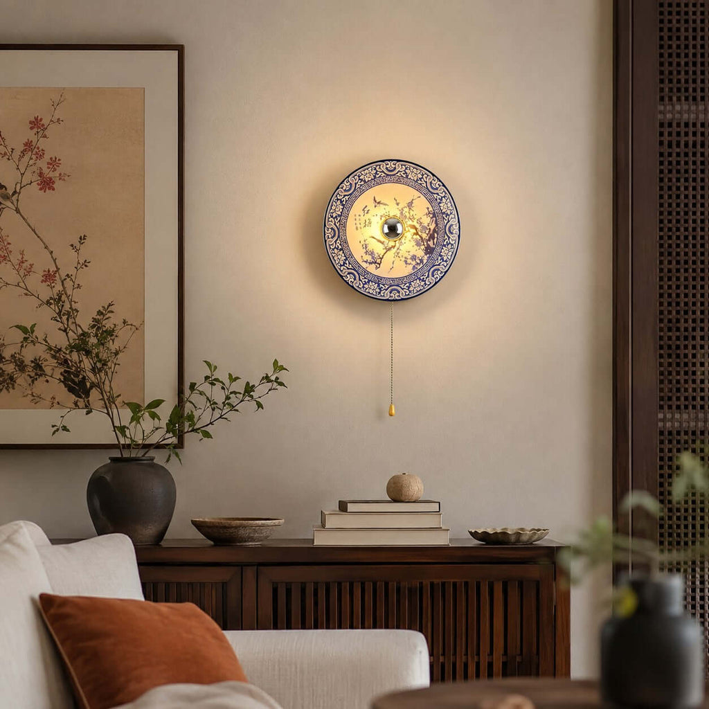 Lit Clérachic porcelain wall light glowing softly above a dark wood sideboard, with a rust cushion on a cream sofa below.