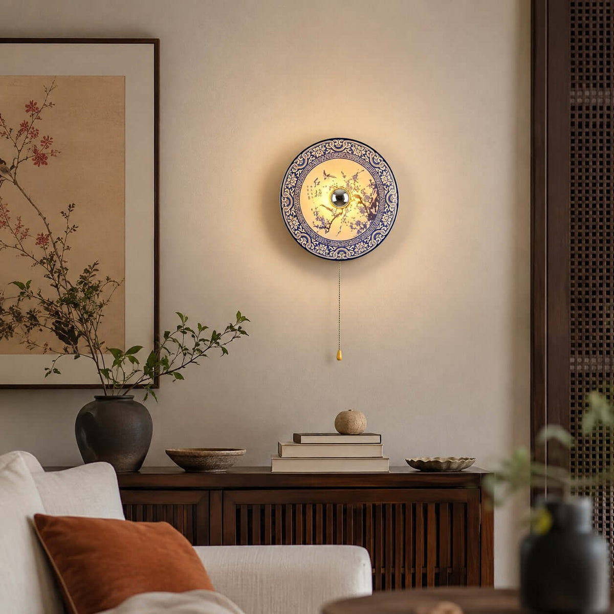 Lit Clérachic porcelain wall light glowing softly above a dark wood sideboard, with a rust cushion on a cream sofa below.