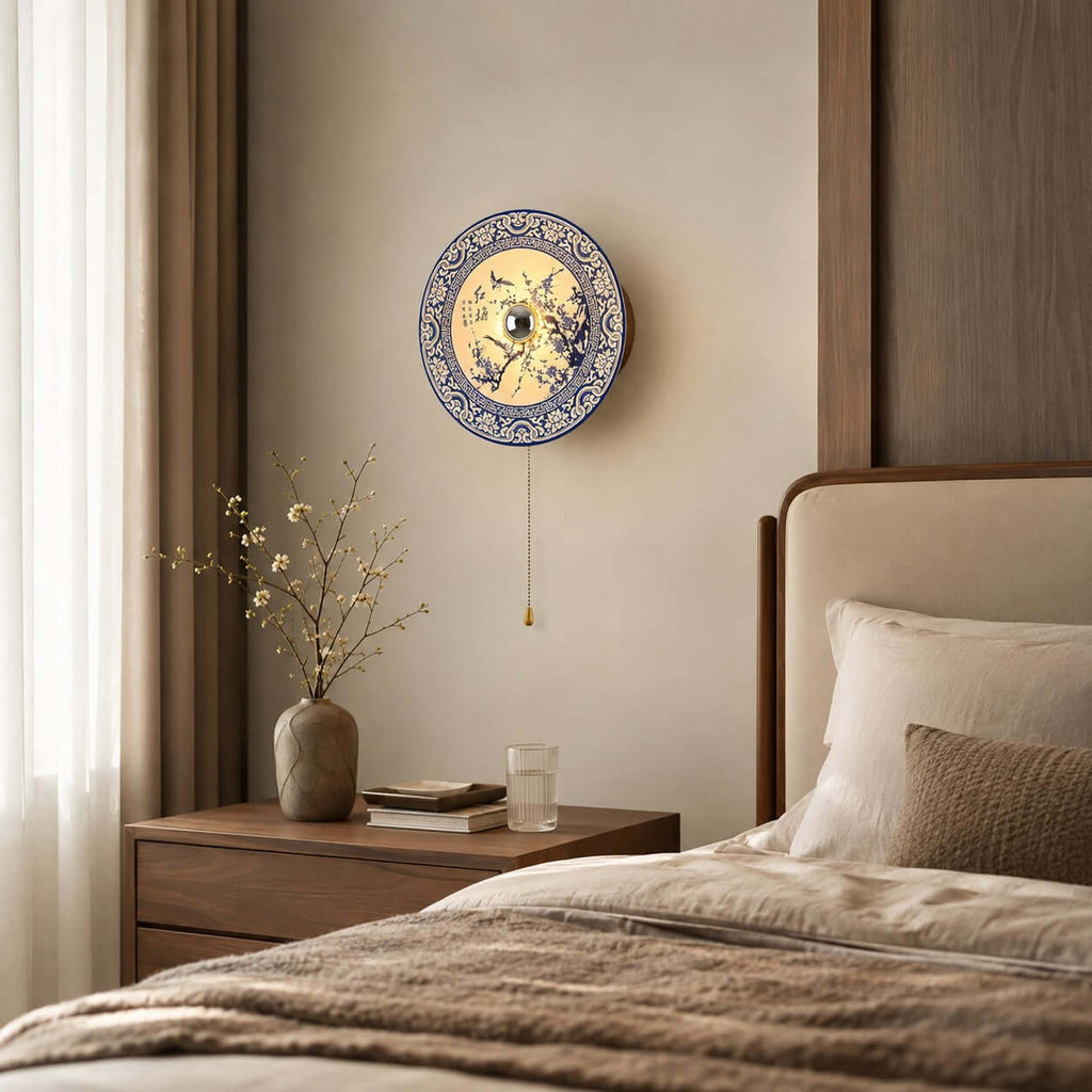 Round Clérachic wall light with a floral blue and white shade, hung over a wood bedside table with a vase of twigs and a glass of water.