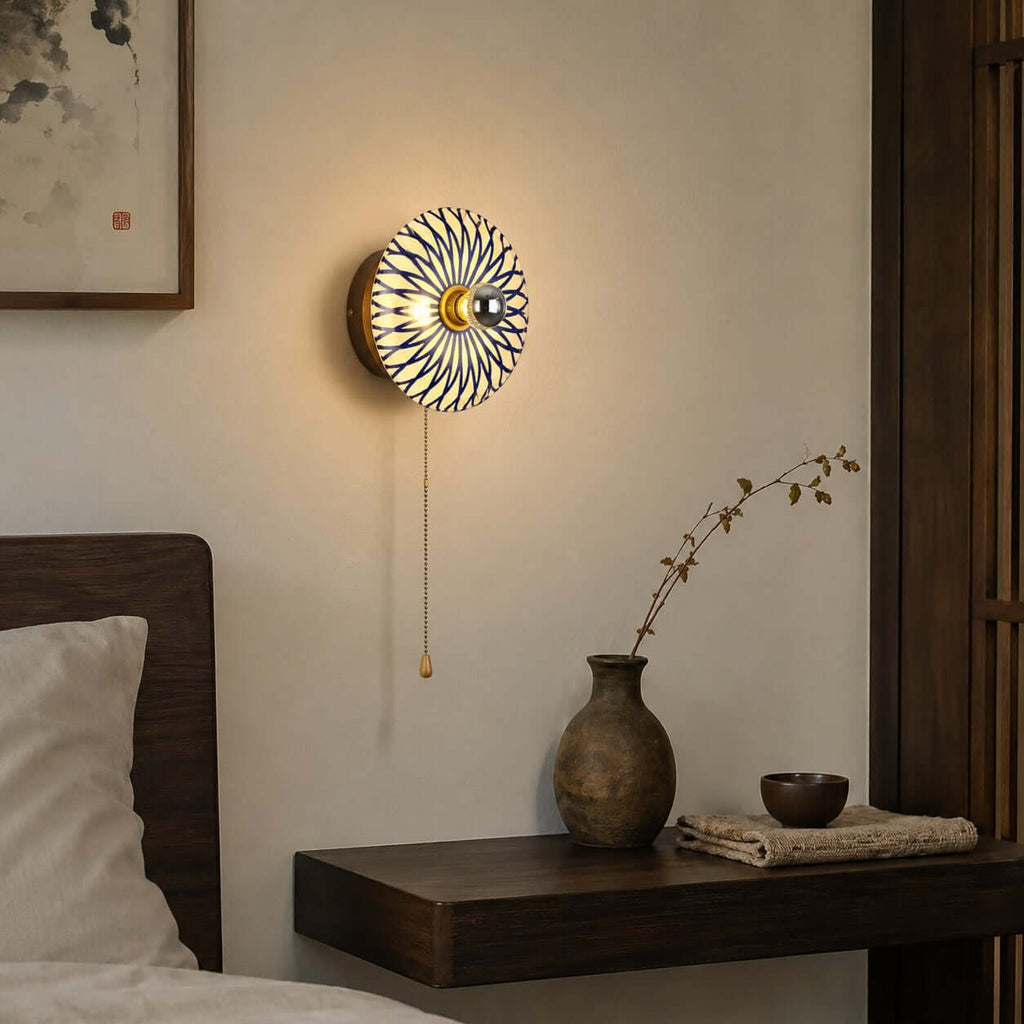 Lit Clérachic wall light with a blue and white petal-pattern shade on a wood backplate, above a floating shelf with a stoneware vase.
