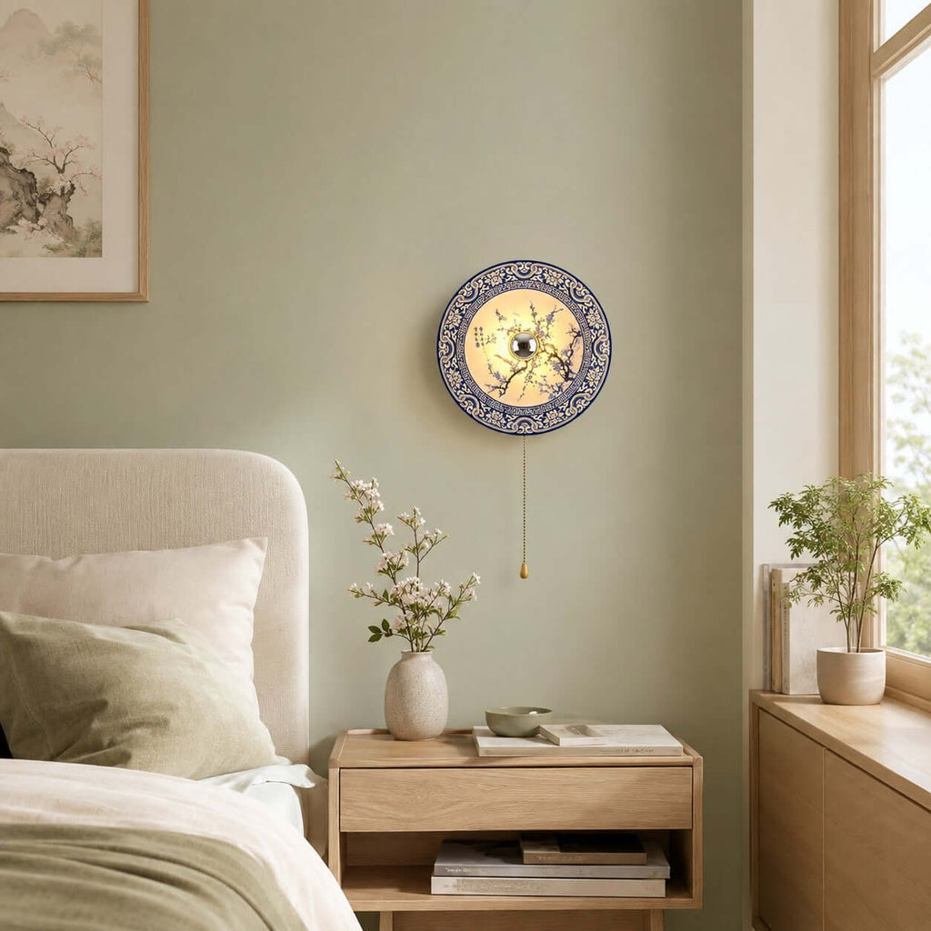 Blue and white Clérachic wall light on a sage-green wall, above a pale bedside table with a vase of blossom and a stack of books.