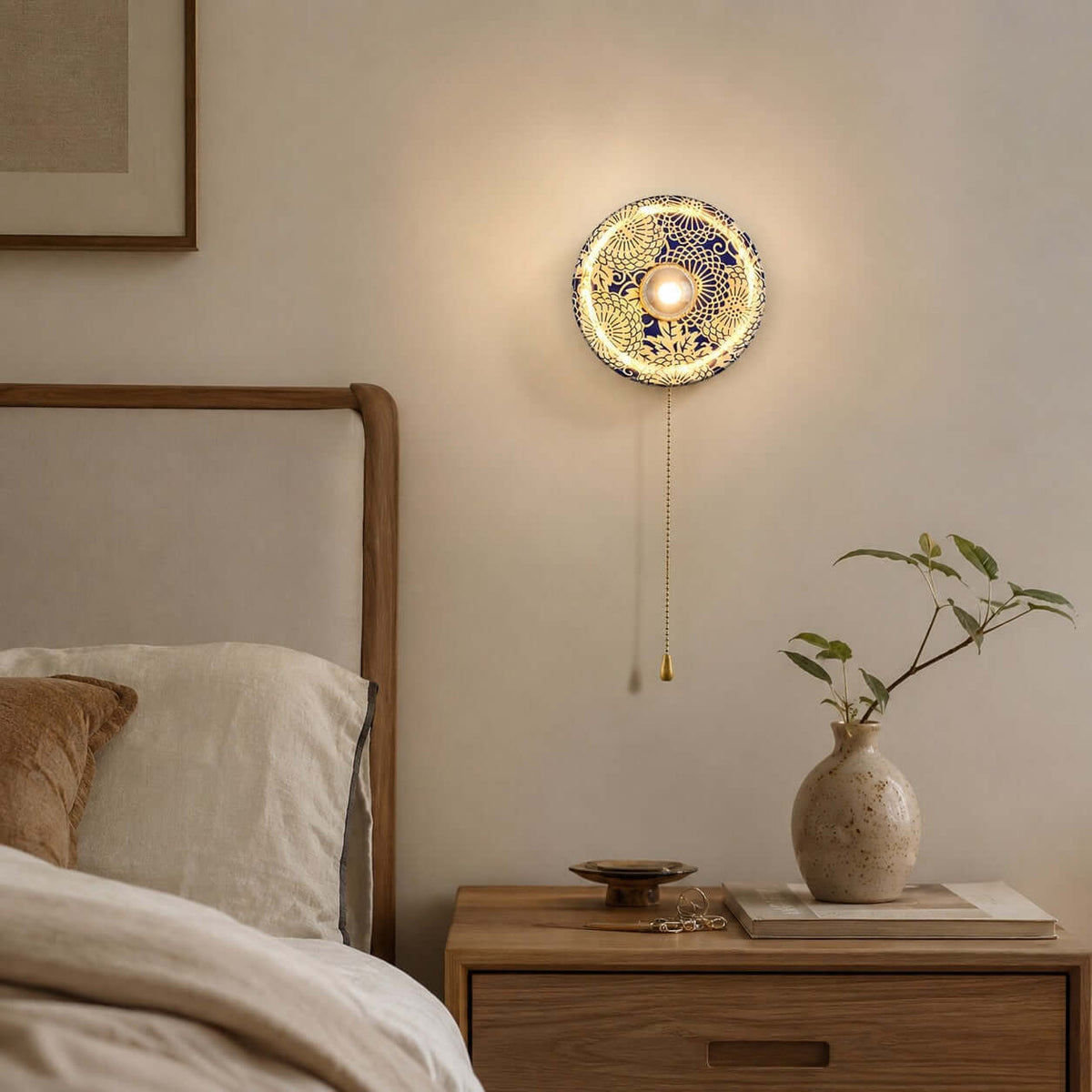 Lit Clérachic wall light with a lacy blue and white shade, above a pale wood bedside table with a vase of green stems.