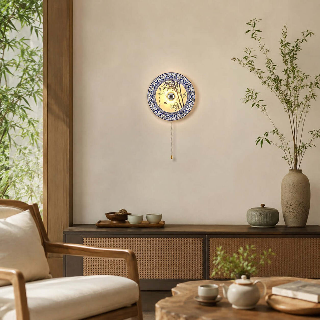 Clérachic wall light with a bamboo design, lit on a plain wall above a woven-front sideboard, with tall green stems in a vase to the right.