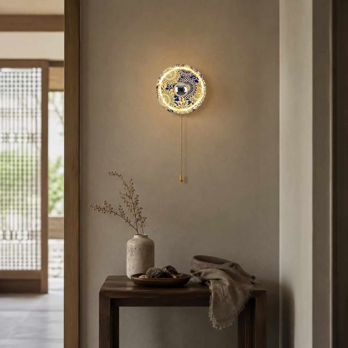 Lit Clérachic wall light with an intricate lattice shade on a tan wall, above a wooden side table with a vase and a folded throw.