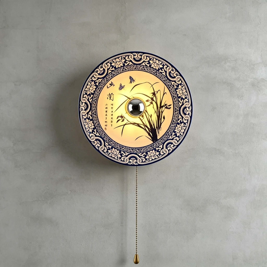 Large lit Clérachic wall light with an orchid design and wide blue border, its brass bead chain hanging beneath on a grey wall.