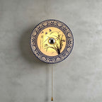 Large lit Clérachic wall light with an orchid design and wide blue border, its brass bead chain hanging beneath on a grey wall.