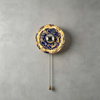 Smaller Clérachic wall light with a dark blue and gold cloud-swirl shade, lit, with a pull chain on a grey wall.