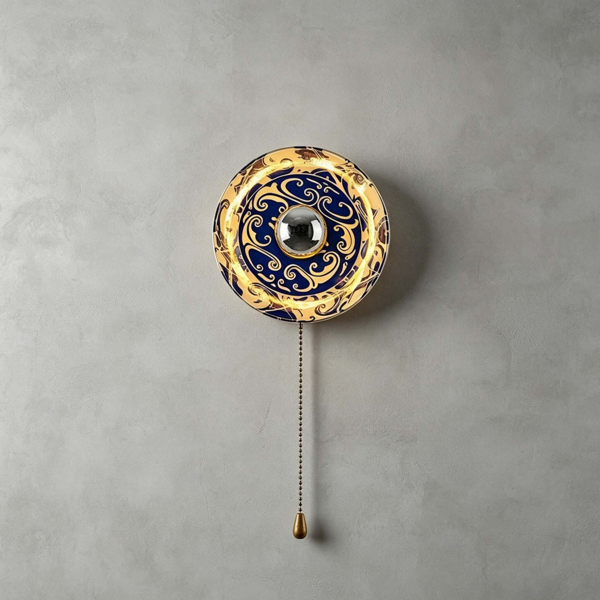Smaller Clérachic wall light with a dark blue and gold cloud-swirl shade, lit, with a pull chain on a grey wall.