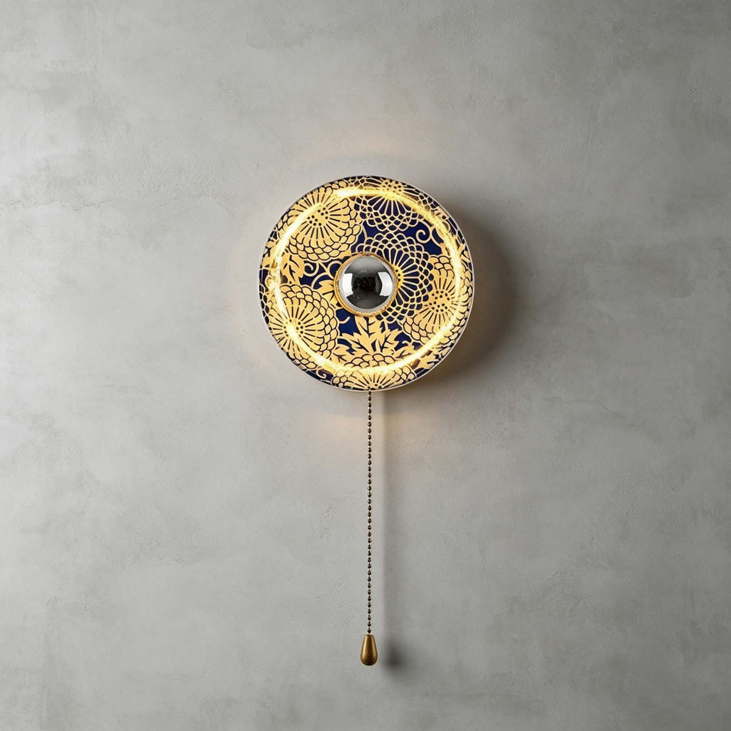 Clérachic wall light with a gold and blue chrysanthemum-lattice shade, lit with a halo of light on a grey wall.