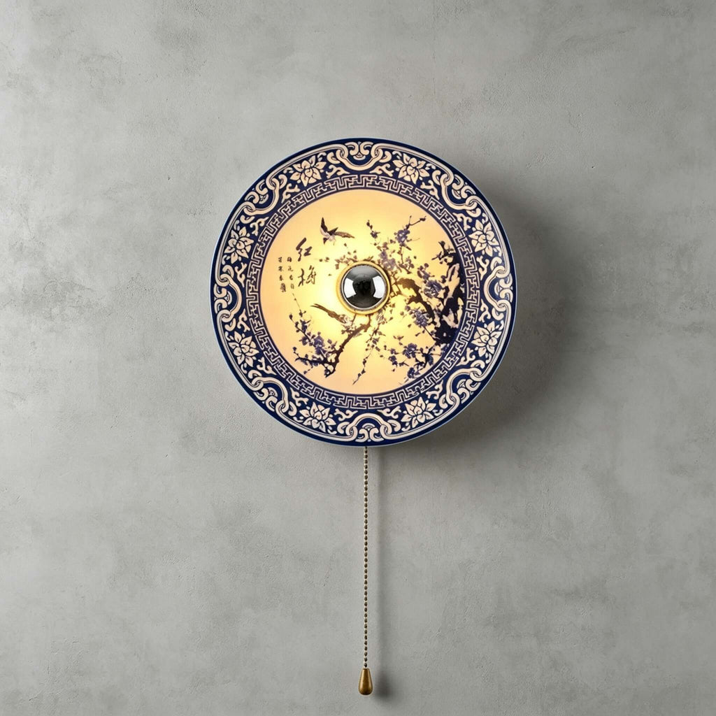 Lit Clérachic wall light with a blossom-branch design and a patterned blue border, its pull chain hanging on a grey wall.