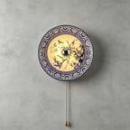 Lit Clérachic wall light with a blossom-branch design and a patterned blue border, its pull chain hanging on a grey wall.